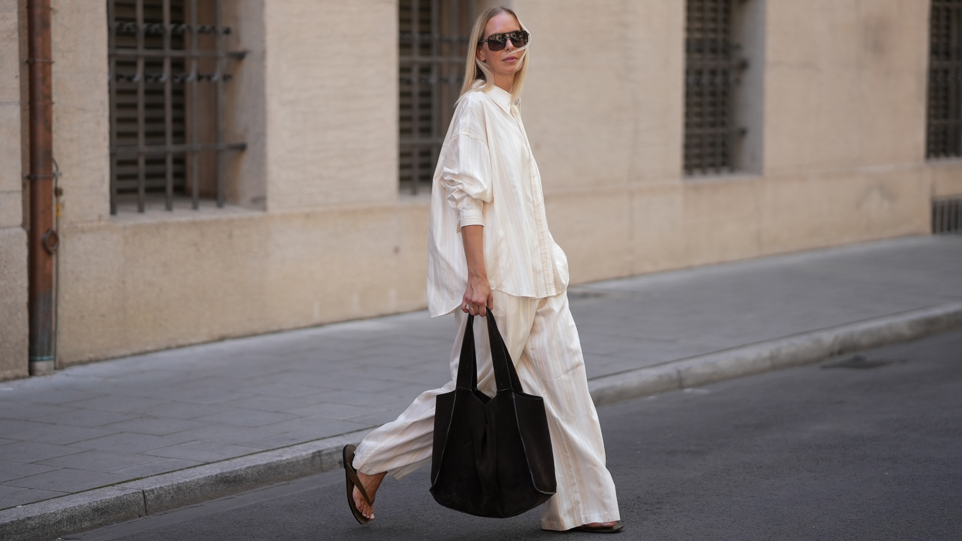 Mirja Klein is seen wearing a matching set consisting of a white striped oversized dobby-weave shirt with collar and buttons down in the front from H&M and a white striped straight pull-on trousers with loose fit and straight legs from H&M; a black suede tote bag with slouchy design and open top from COS; black oversized aviator sunglasses from Tom Ford; a pair of khaki green suede flip flops from A.EMERY on June 18, 2025 in Munich, Germany.