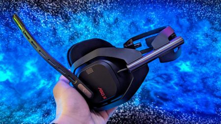 A person holds a black gaming headset with a prominent microphone against a vibrant blue and black galaxy-like background, creating a futuristic feel.