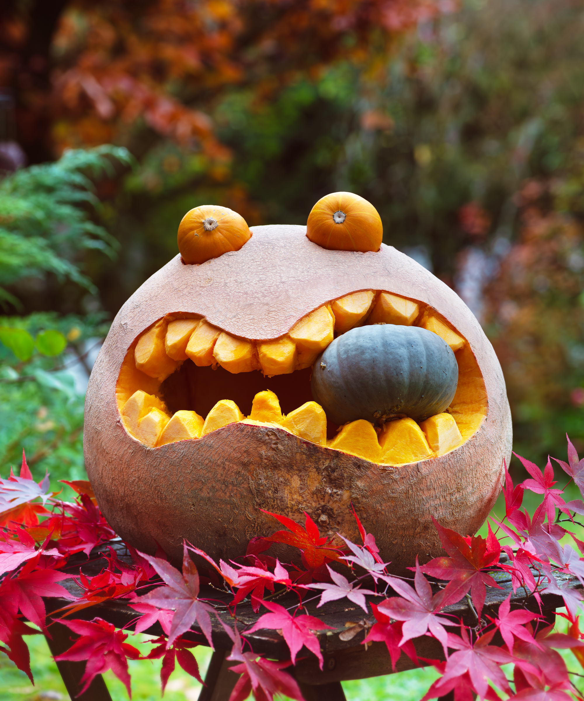 Funny Halloween pumpkin carving of a round monster with big jagged teeth, tiny pumpkins as eyes, and a green gourd inside its mouth