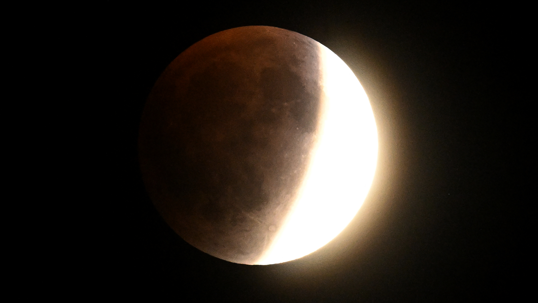 A full moon is chown with Earth's crescent shadow falling upon it during a total lunar eclipse. The extreme right edge is bathed in direct sunlight while the left is bathed in a rusty shadow.