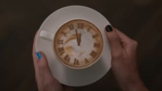 Taylor Swift's hands with blue and black nails holding a coffee cup with latte art of a clock that is about to strike midnight.