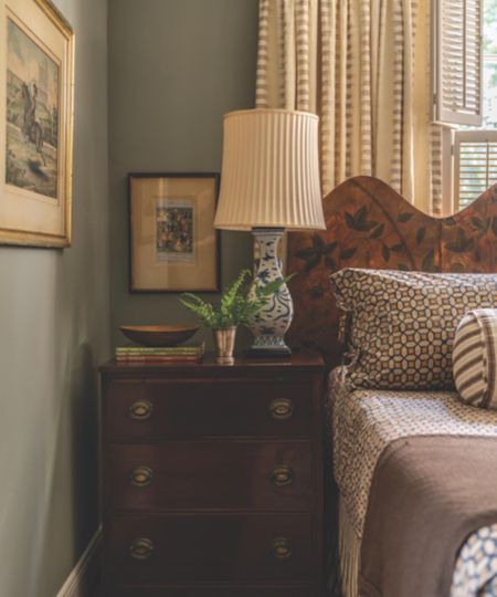 Bedroom with pale blue walls, a mahogany nightstand, a screen headboard, vintage china lamp, patterned geometric bedding, and antique prints on the walls.