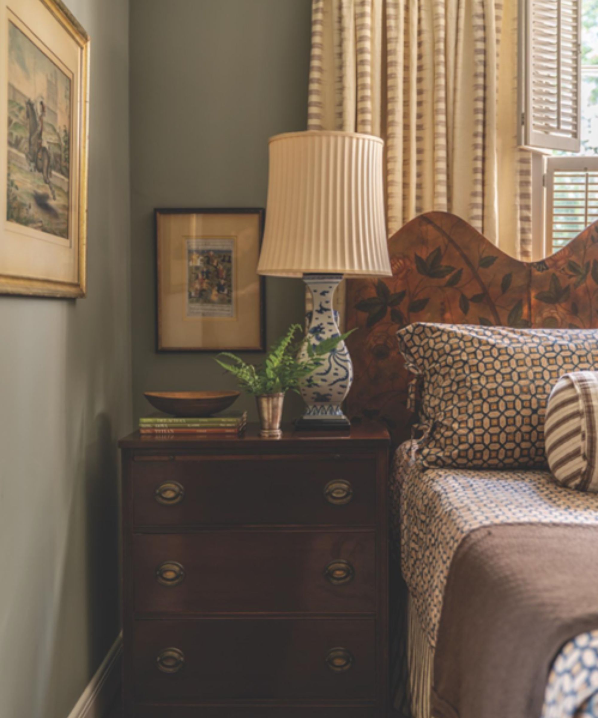 Bedroom with pale blue walls, a mahogany nightstand, a screen headboard, vintage china lamp, patterned geometric bedding, and antique prints on the walls.