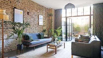 Living area of open-plan space with exposed brick wall, concrete floor covered with large jute rug, Crittall-style French doors and mid-century furniture including two sofas and a coffee table