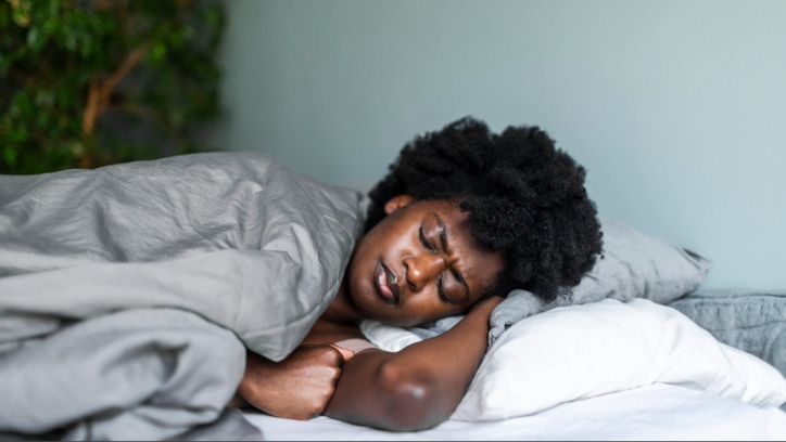 A woman with an afro sleeping on her side covered in grey duvet while she sweats in her sleep, thinking of a cooling mattress.