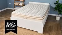 The Saatva Classic mattress in a blue bedroom and placed on a white wooden bedframe with a Black Friday sales badge overlaid on the bottom left hand corner