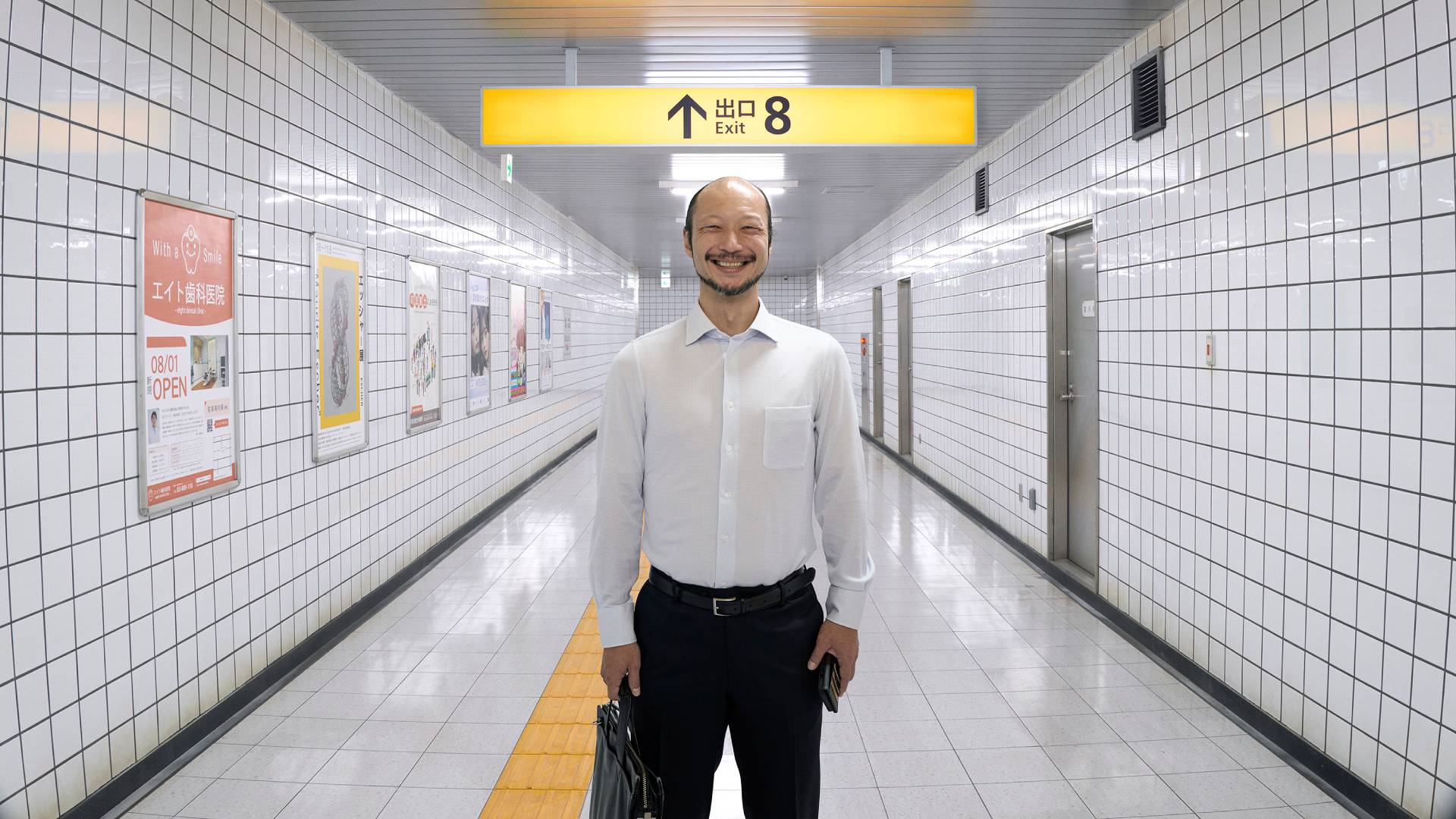 An image from Exit 8 showing a clean, bright passageway of a Japanese underground metro with a single suited man standing and smiling