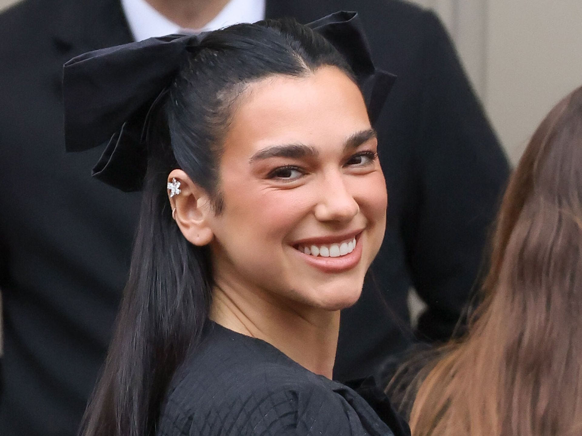 Dua Lipa smiles at the camera wearing a black hair bow