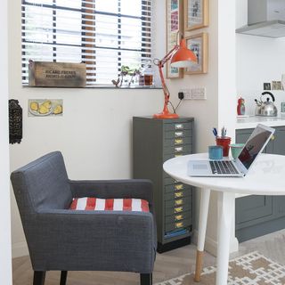 white and grey open plan office and kitchen, with a grey chair with red and white striped cushion next to a white circular table with wood dipped legs, and a tall and skinny grey filing cabinet with an orange lamp on top
