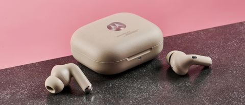The Motorola Moto Buds+ earphones are lying on a dark surface either side of their charging case, pictured in front of a pink background.