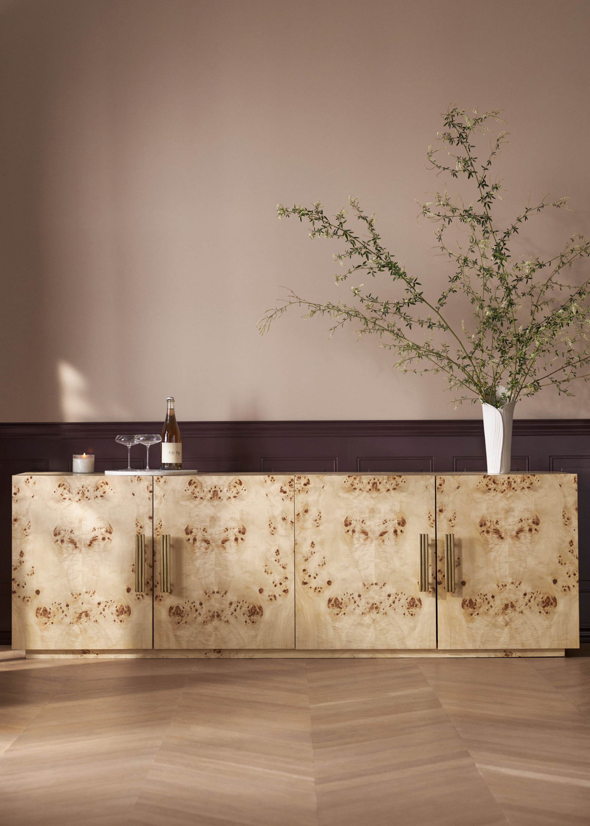 Erin Fetherston Exclusively for Anthropologie burl wood credenza with large foliage on top set in front of a dusty pink wall with a dark colored paneling running along the bottom third edge and herringbone wood flooring