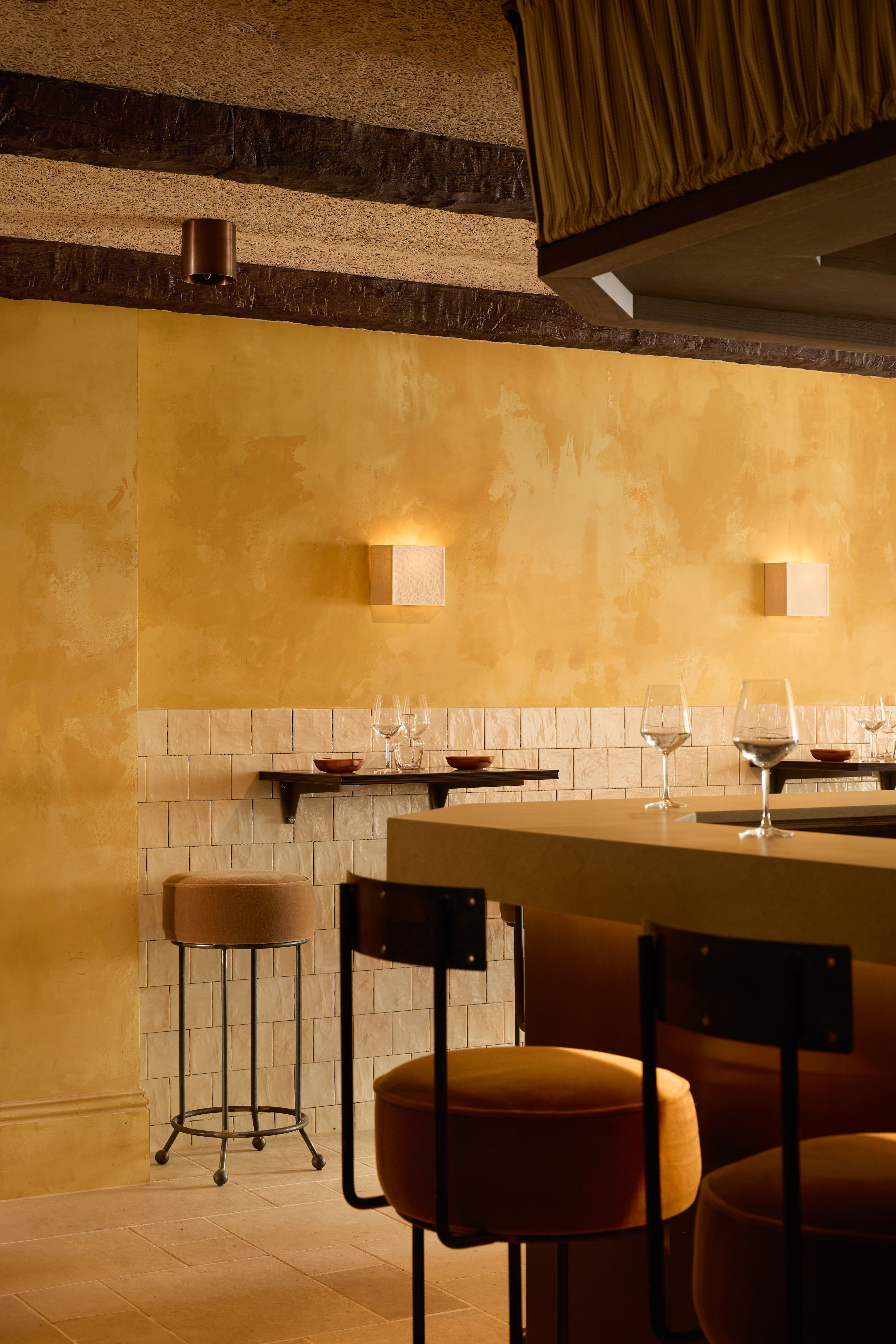 A warmly furnished restaurant with butter and ochre plaster walls, wrought iron chairs with neutral cushions, statement ceilings in brick or plaster relif, and stripy furniture paired with plants.
