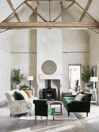 living room with high vaulted ceiling, beams, textured walls, log burner, consoles, green velvet sofa and chair, floral sofa, lime washed floorboards, rug, coffee table