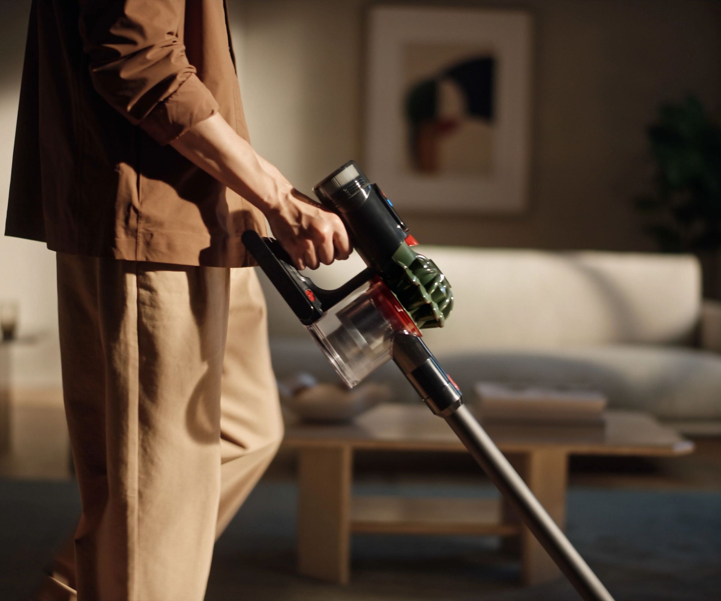 Person in brown and tan clothing holding Dyson V8 Cyclone in green and gray colorway in front of blurred-out living room.