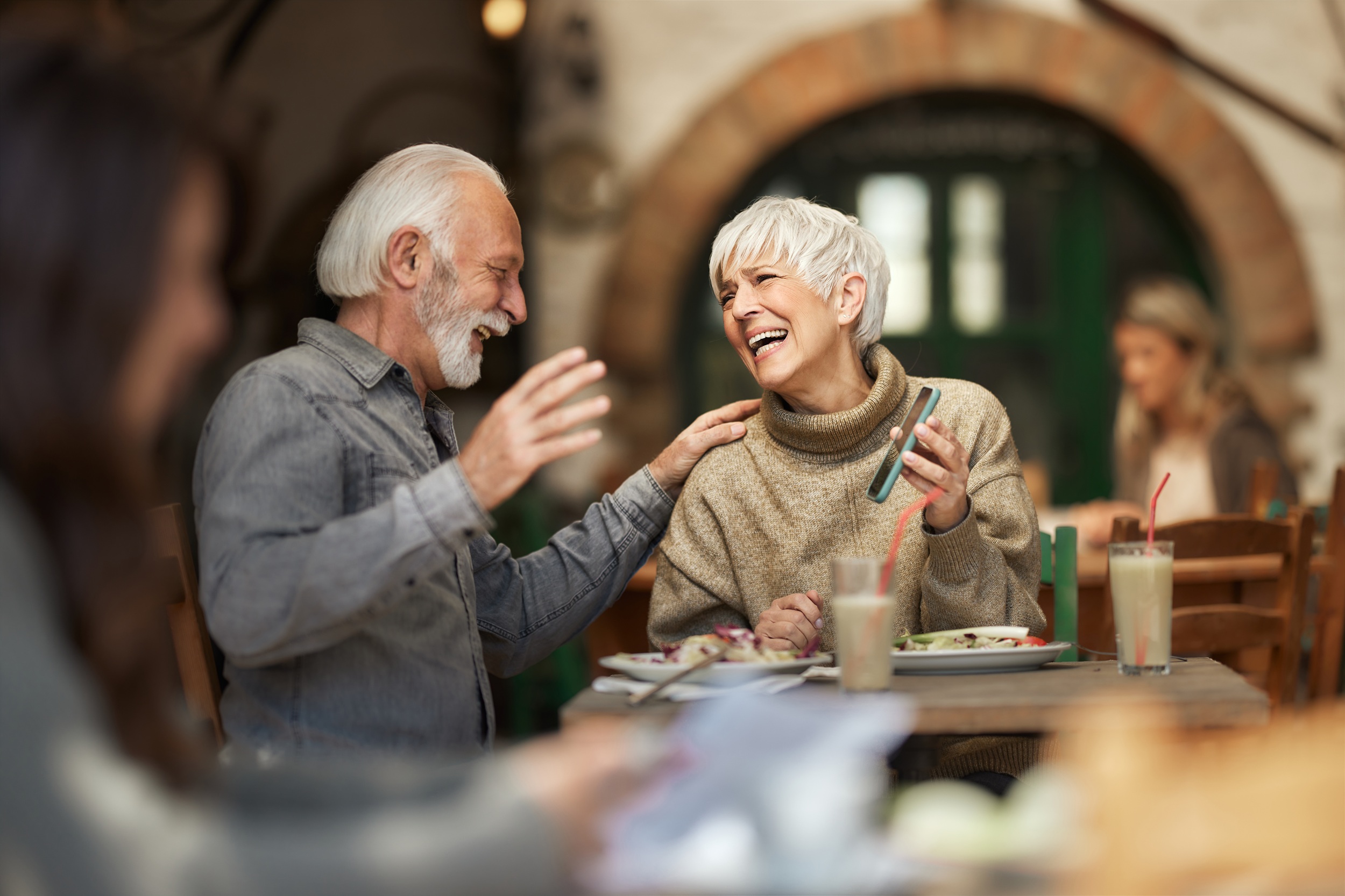 Cheerful senior woman having fun while showing her husband a funny text message on her cell phone during a meal in a restaurant.