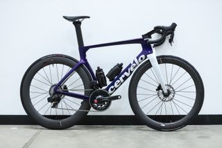 A blue and silver Cervelo S5 stands against a white wall