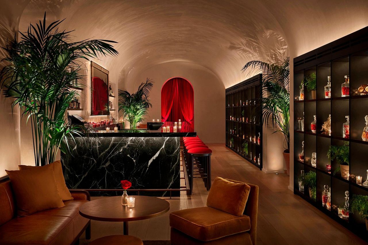 A superbly decorated, hyper-modern hotel features minimalist cozy furniture including neutrally-hued large couches, standing lamps, tiled floors, cream curtains, wall-to-wall windows, and breathtaking views of the city.