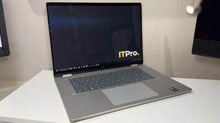 A gray laptop sits open on a white desk, its dark screen displaying the ITPro logo. A clean, modern office setting feels quiet and professional.