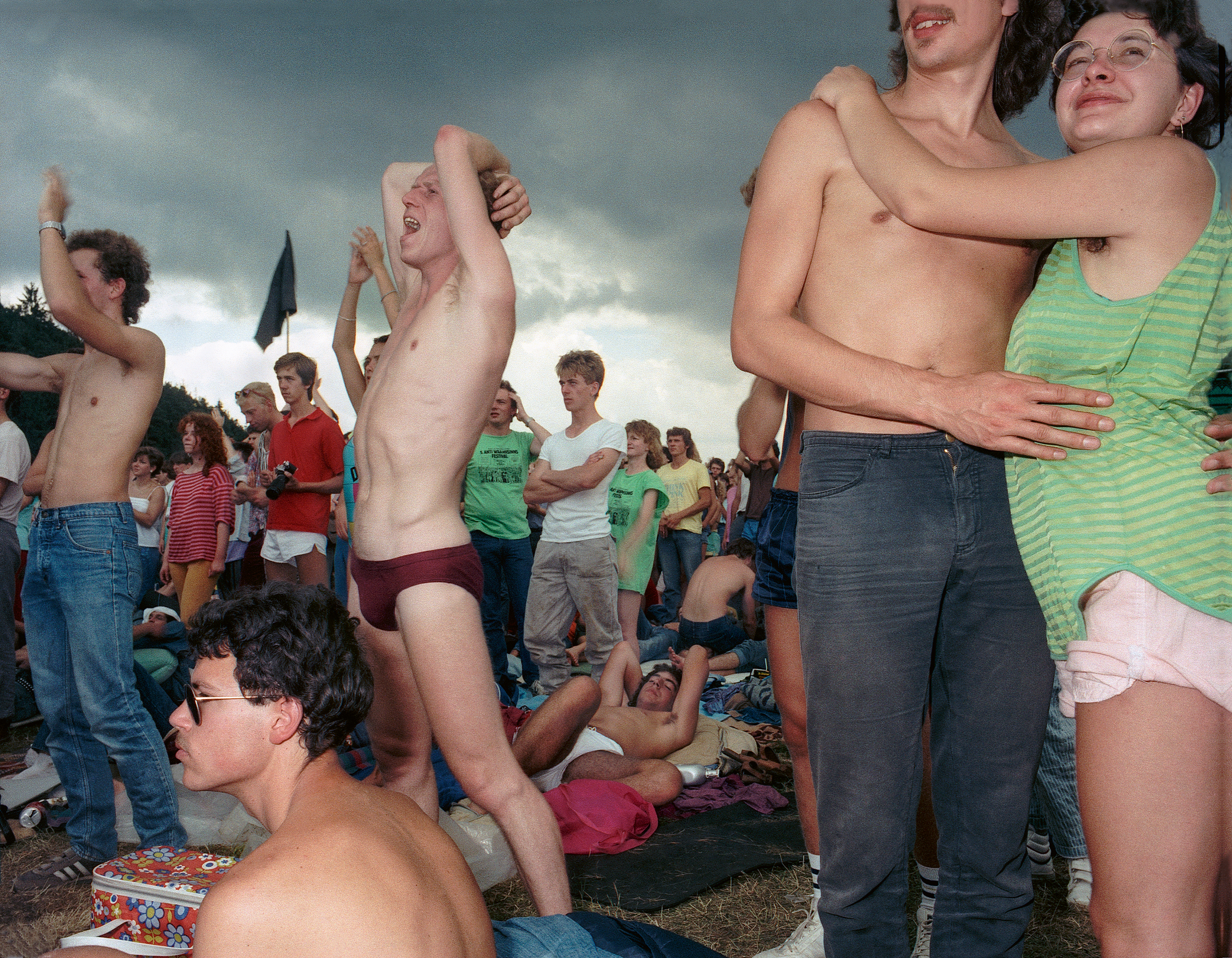 At the 1986 Wackersdorf anti-nuclear protest, a shirtless man raises his arm mid-shout as a couple embrace beside him, a dark storm-clouded sky filling the background.