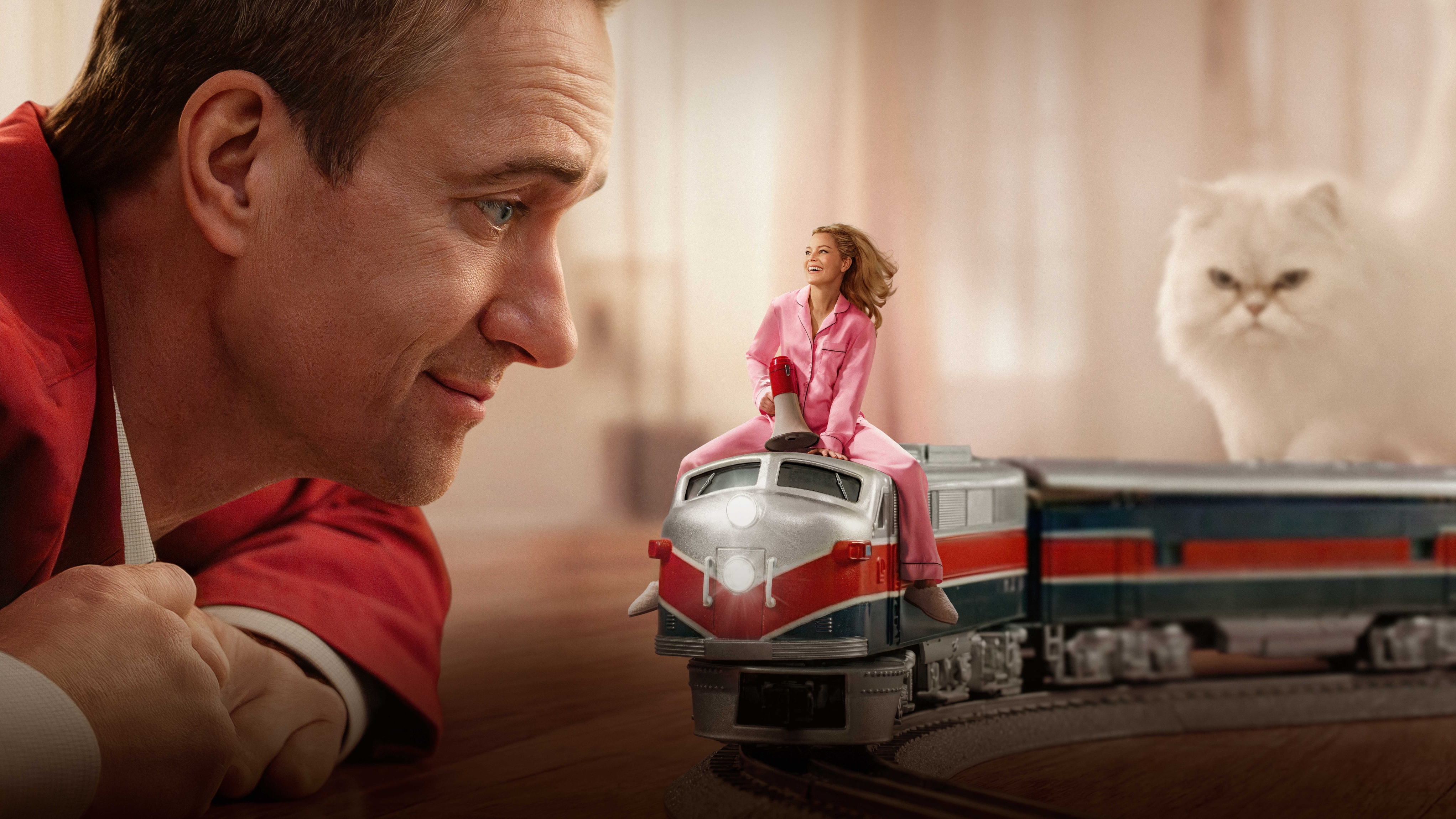 Les (Matthew Macfadyen) looks at his miniature wife Lindy (Elizabeth Banks) sitting on a toy train with a fluffy white cat behind them.