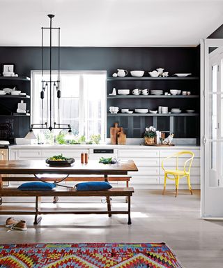 An example of kitchen shelving ideas showing an open plan kitchen with white cabinets, dark walls and a wooden dining bench