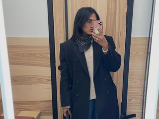 Chiara wears a long black coat, white sweater, jeans, and neckerchief in a mirror selfie