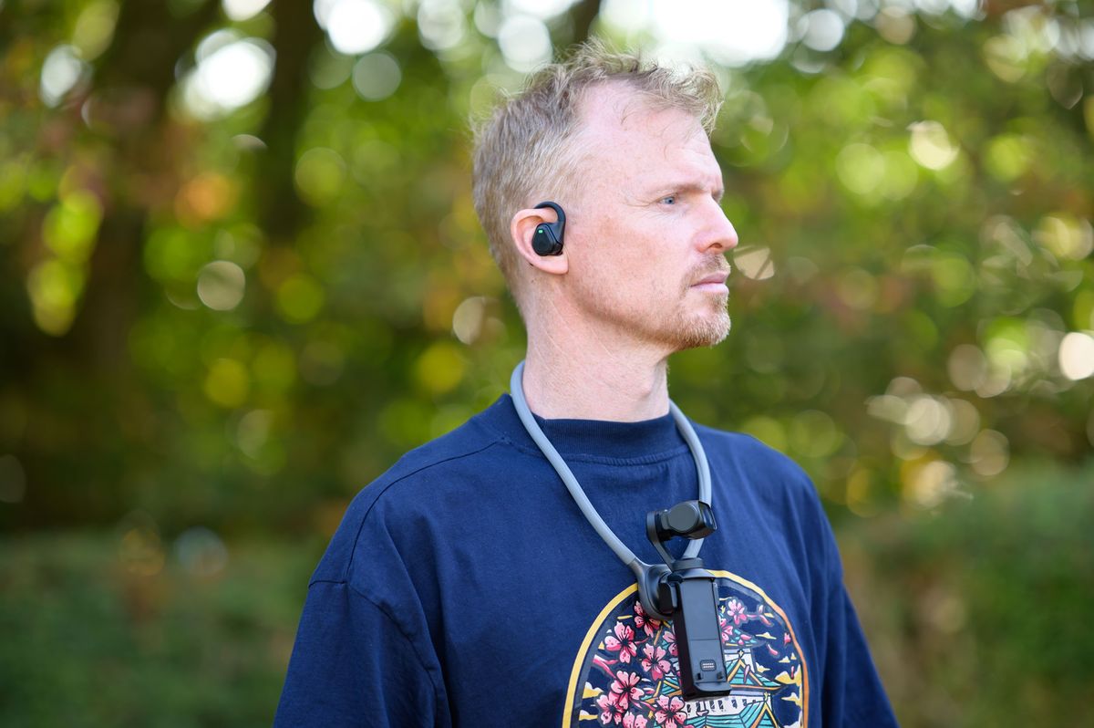 Tim Coleman wearing the Insta360 POV Head Tracker accessory in his ear, and Luna Ultra camera on his chest attached to a neck mount, outdoors with dappled light in background foliage