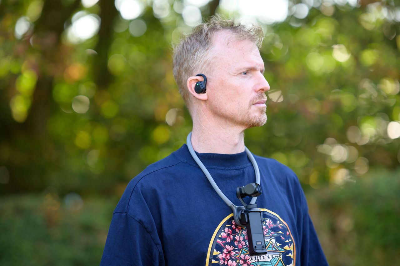 Tim Coleman wearing the Insta360 POV Head Tracker accessory in his ear, and Luna Ultra camera on his chest attached to a neck mount, outdoors with dappled light in background foliage