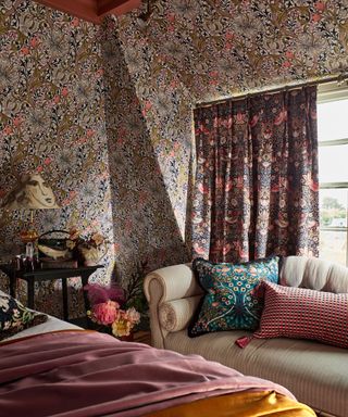 Floral, maximalist, bold bedroom with patterned curtains, wallpaper, soft furnishings, bed dressed with blankets and cushions, cream striped sofa with cushions beside window