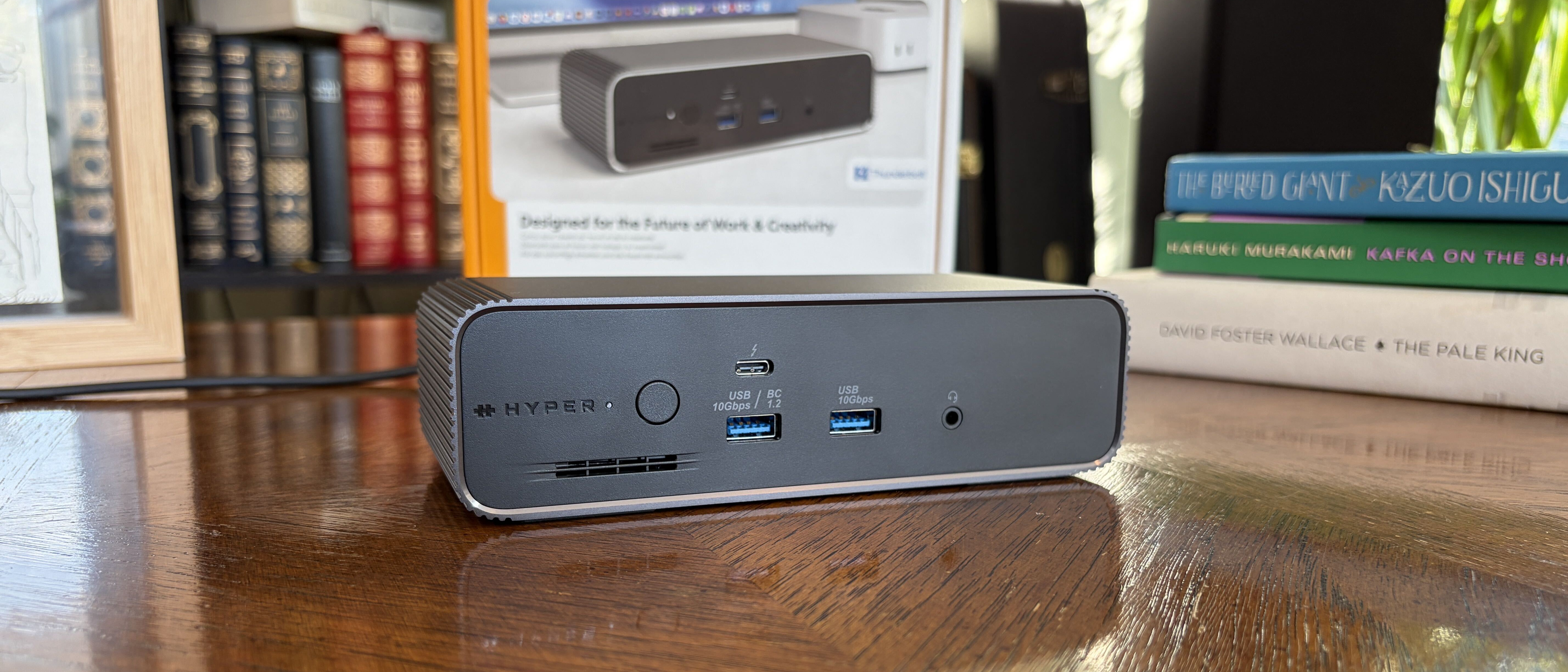 A gray HyperDrive Next docking station with multiple ports displayed on a wooden desk, surrounded by colorful books and a framed image.