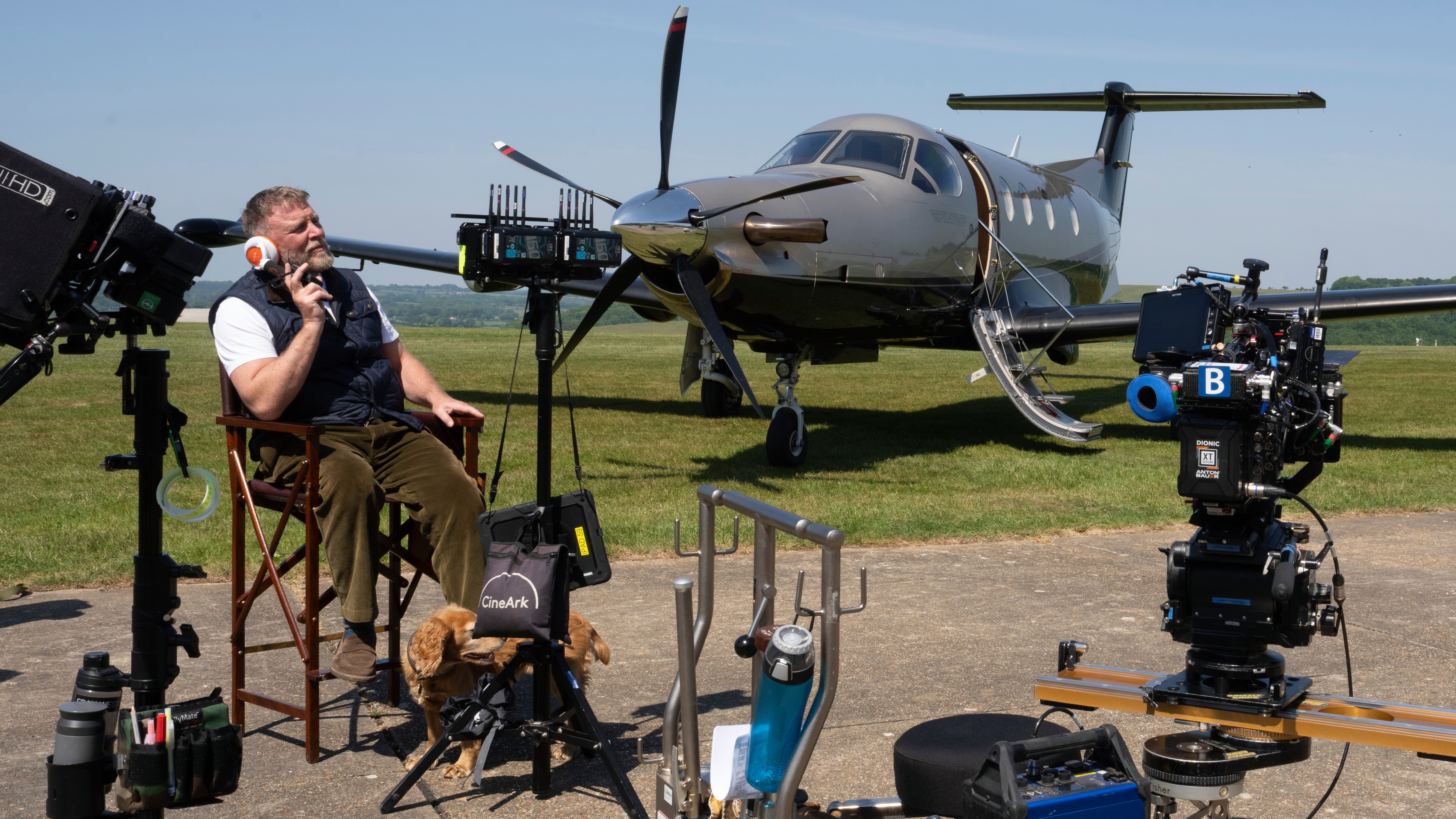 A behind the scenes photo Netflix released from the set of "The Gentlemen" season 2 showing Guy Ritchie sitting in a director's chair surrounded by film equipment, with a plane in the background