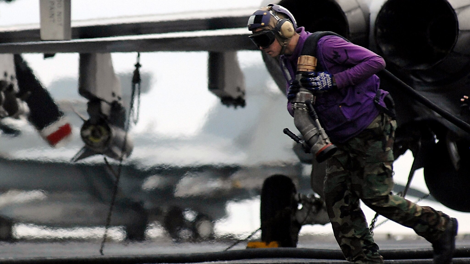 A man wearing a pilot's helmet and sunglasses and a purple jacket and camouflage pants carries a large hose away from a plane. 