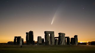 stonehenge stands with a glowing sky in behind. a white comet with large tail is visible over the stones