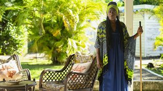 Dorna Bray (Judith Jacob) stands out on her veranda next to some garden chairs. She is holding a broom and looking deep in though. Behind her the garden is lush and green, and there is a white outhouse on the right hand side of the picture.