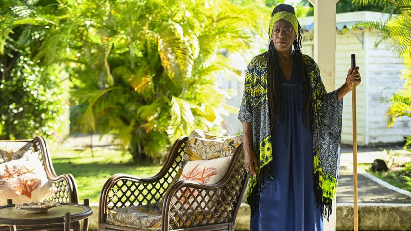 Dorna Bray (Judith Jacob) stands out on her veranda next to some garden chairs. She is holding a broom and looking deep in though. Behind her the garden is lush and green, and there is a white outhouse on the right hand side of the picture.