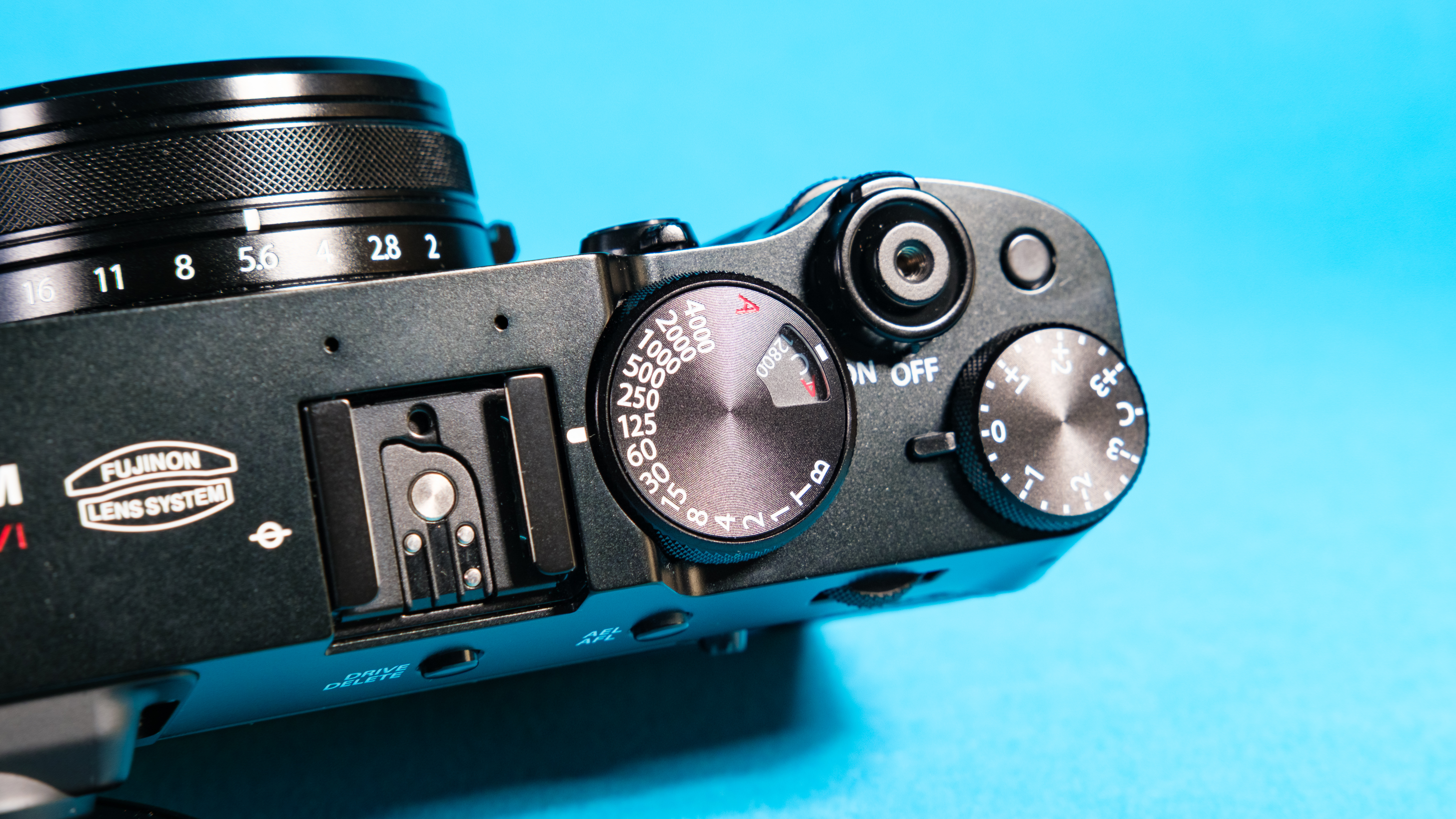 The Fujifilm X100VI mirrorless camera against a blue background lying on its side with the dials showing.