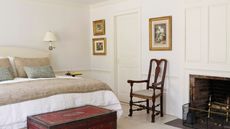 A white panelled bedroom with a large double bed with white bedding, patterned trunk at the end of the bed, wooden chair, exposed brick fireplace, and three artworks in gold frames.