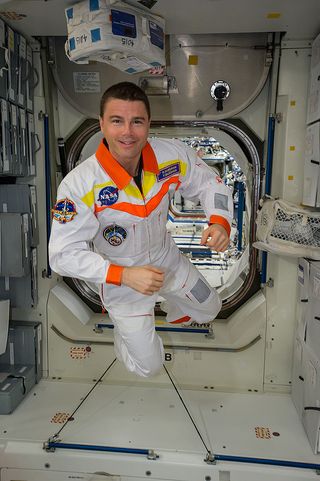 reid wiseman floating beside an entranceway in the international space station, wearing a white and orange jumpsuit
