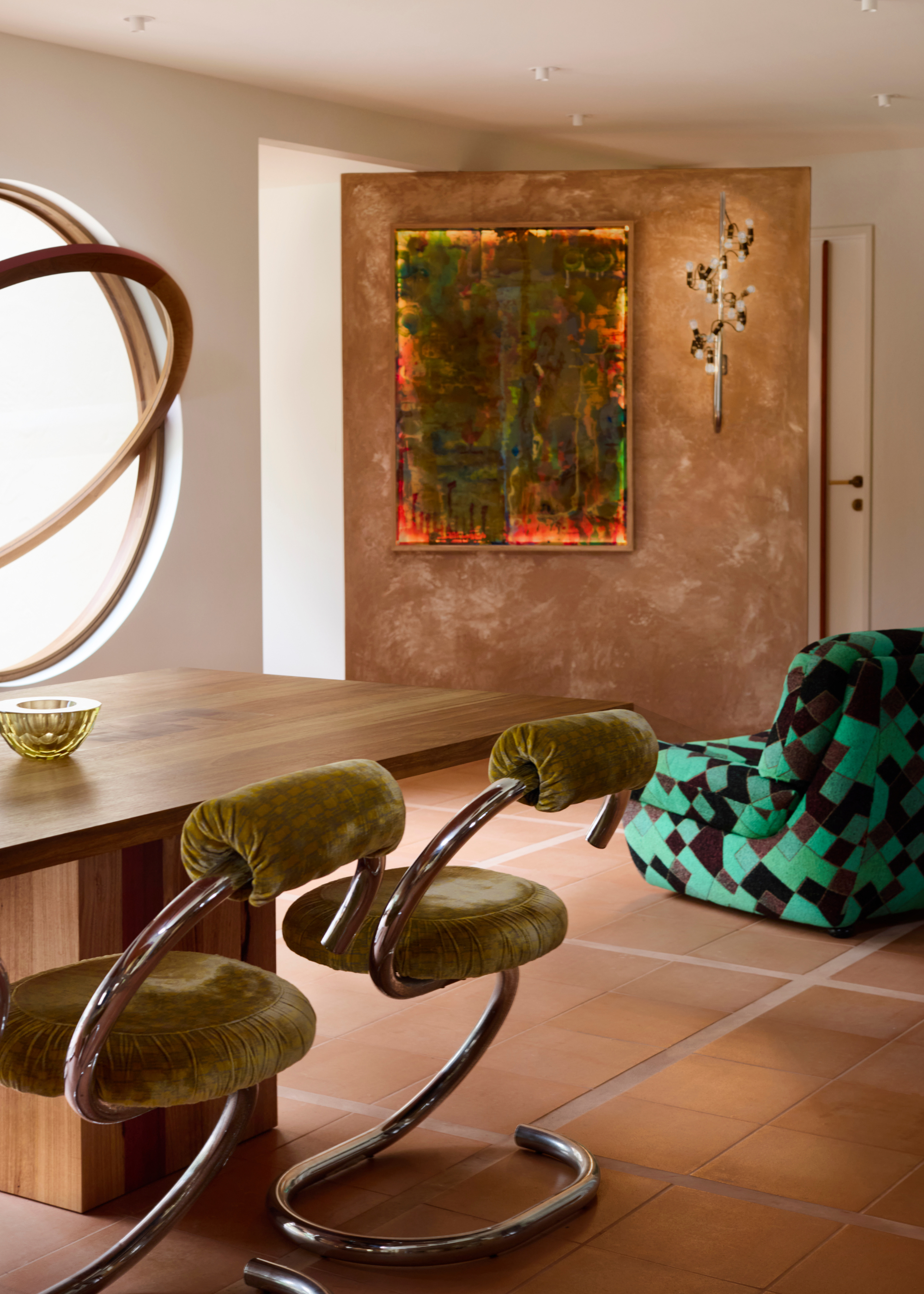 A modern space with a giant wood-framed porthole window that is partly open is next to an angled limewash-style finish wall with artwork and a wall light on it, with a vibrant green checkered lounge chair in front and the bottom end of a wooden dining table with steel framed chairs with green seats and backrests