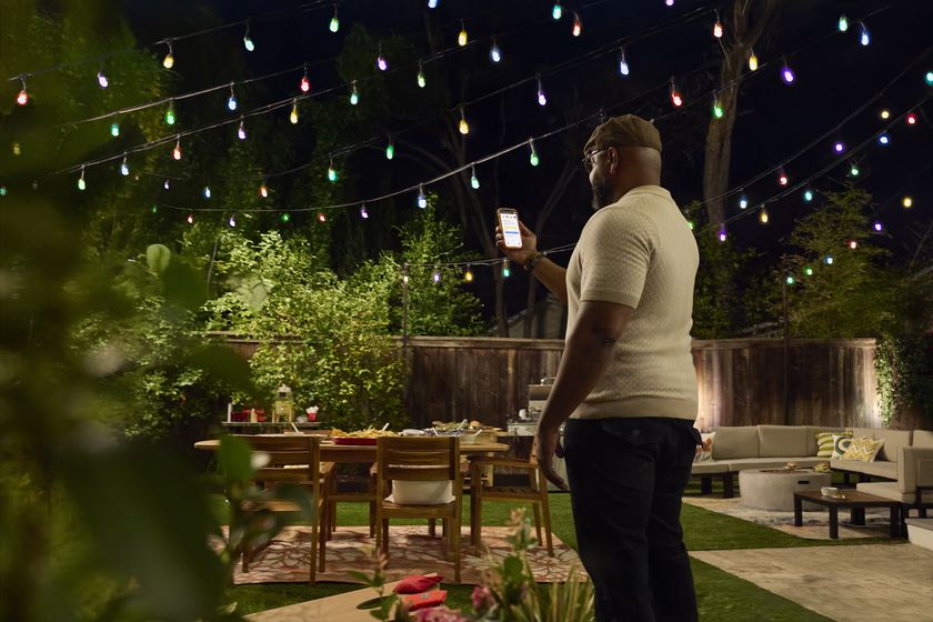A man stands in a fenced yard at night, with multiple multi-colored string lights overhead. The man is adjusting the colors of the lights using the Hubspace app on a mobile phone. 