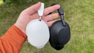 A hand holding the White Sonos Ace and black Sony WH-1000XM5 over-ear headphones by their headbands in front of a patch of grass.