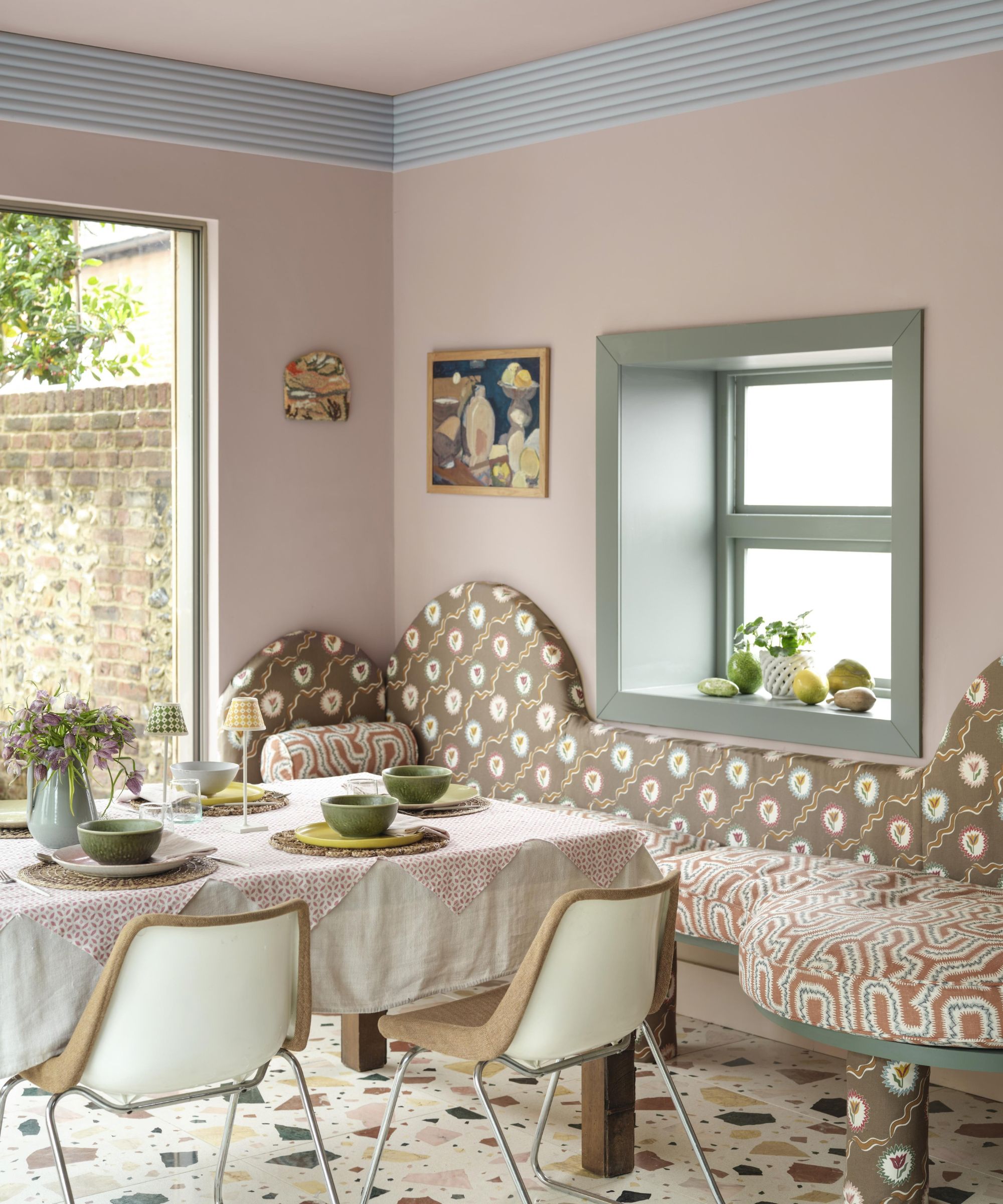 Pale pink dining nook with sage green window frame, a built-in banquette upholstered in brown printed fabric, a dining table with a gingham tablecloth, metal dining chairs, and a terrazzo floor.