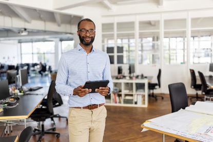 A confident entrepreneur stands with a digital tablet in hand in a modern coworking office, smiling at the camera, representing professionalism and success in a startup.