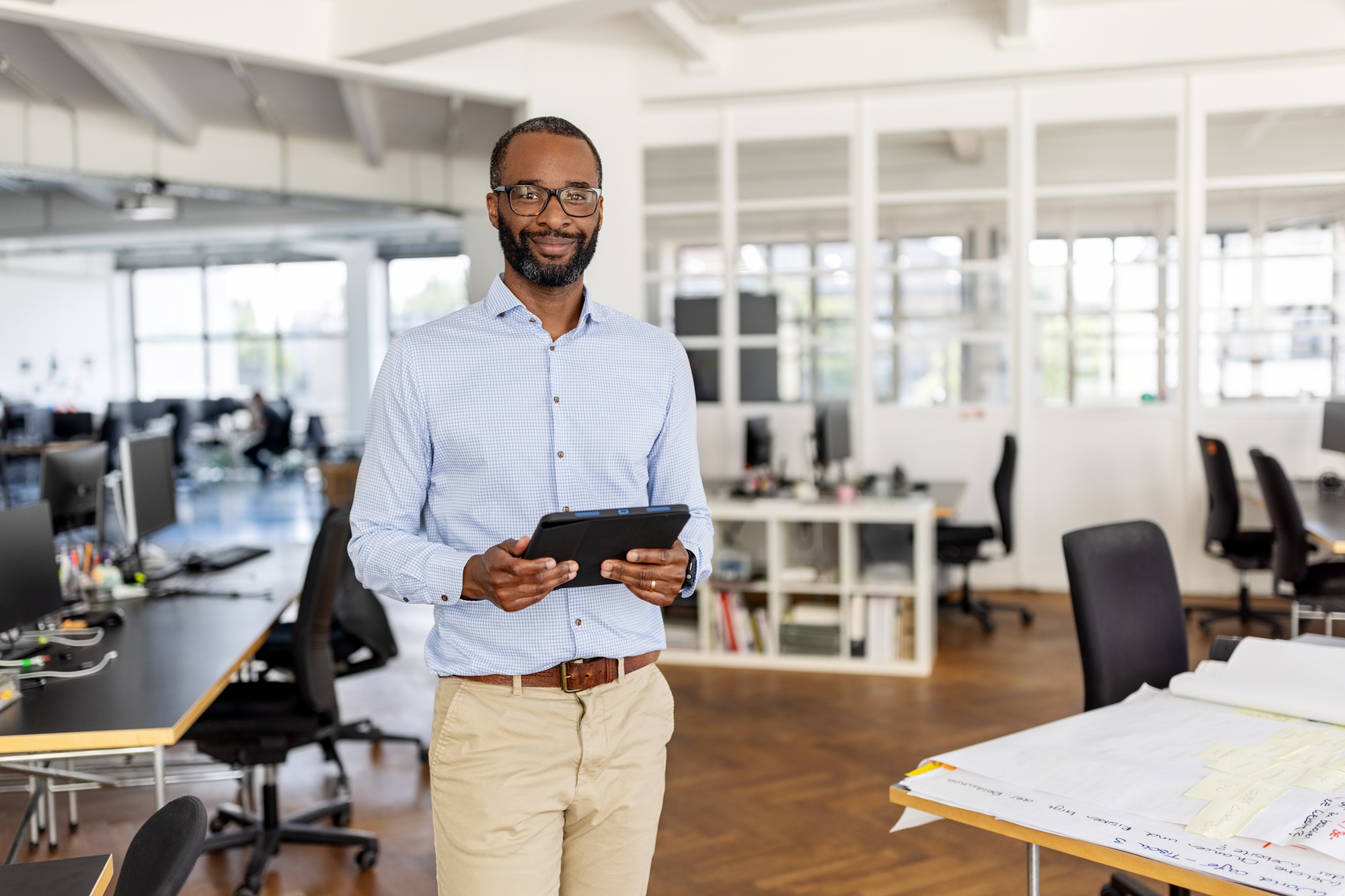 A confident entrepreneur stands with a digital tablet in hand in a modern coworking office, smiling at the camera, representing professionalism and success in a startup.
