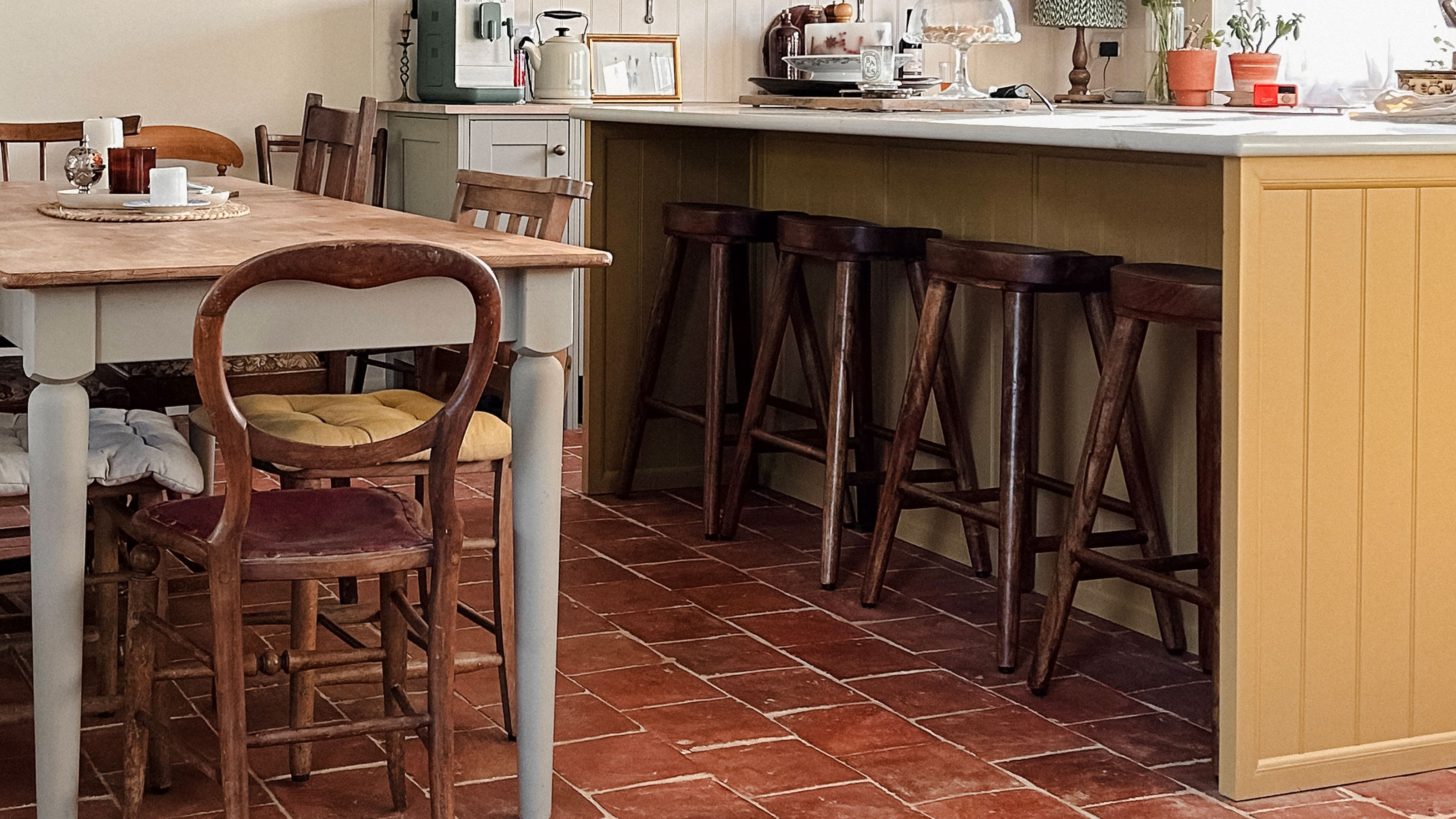 Yellow panelled kitchen island with dark wooden bar stools on tiled terracotta floor beside kitchen table