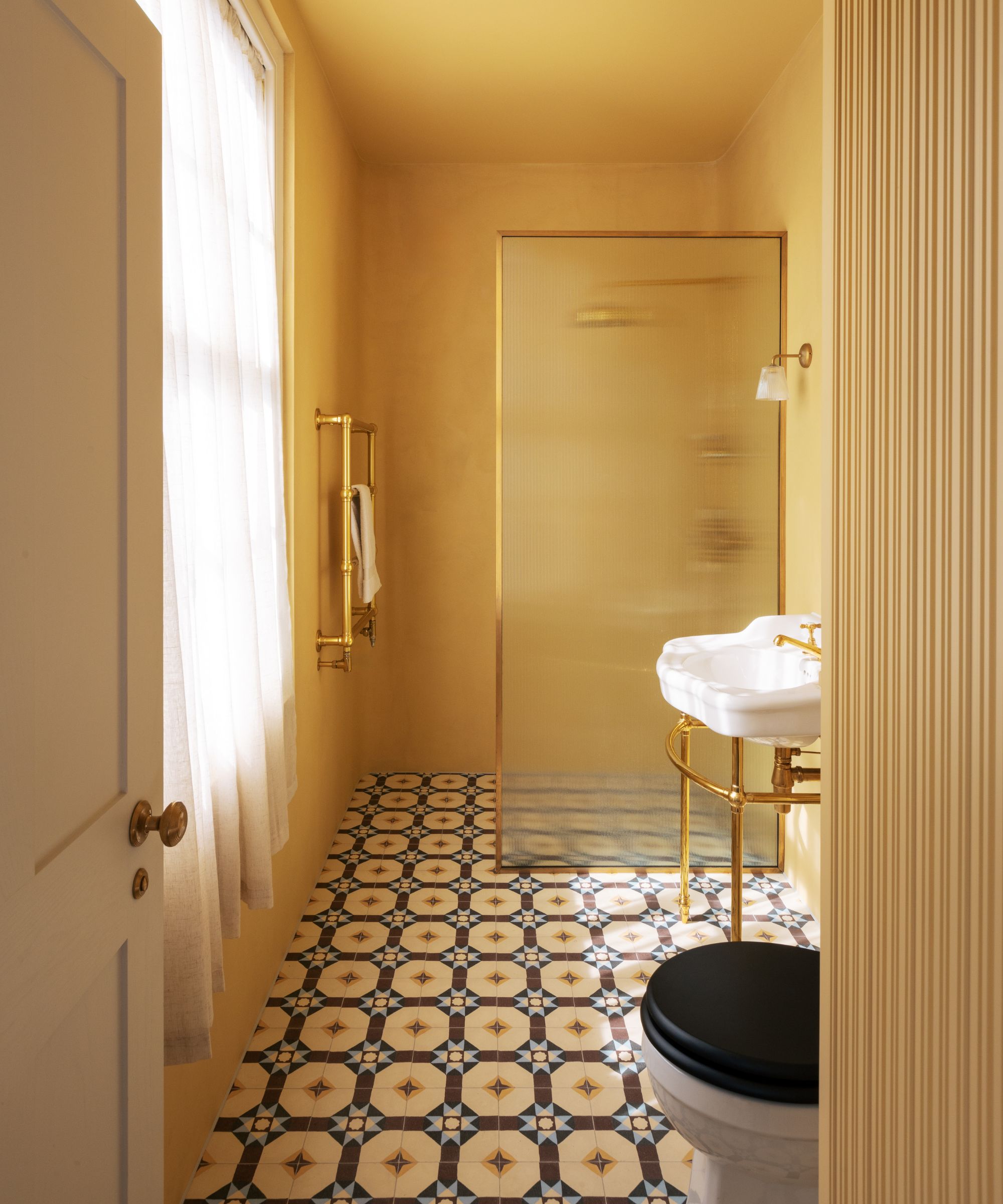 A narrow yellow-painted bathroom with bold geometric patterned floor tiles, a brass console basin, frosted glass shower screen and a black-lidded WC.