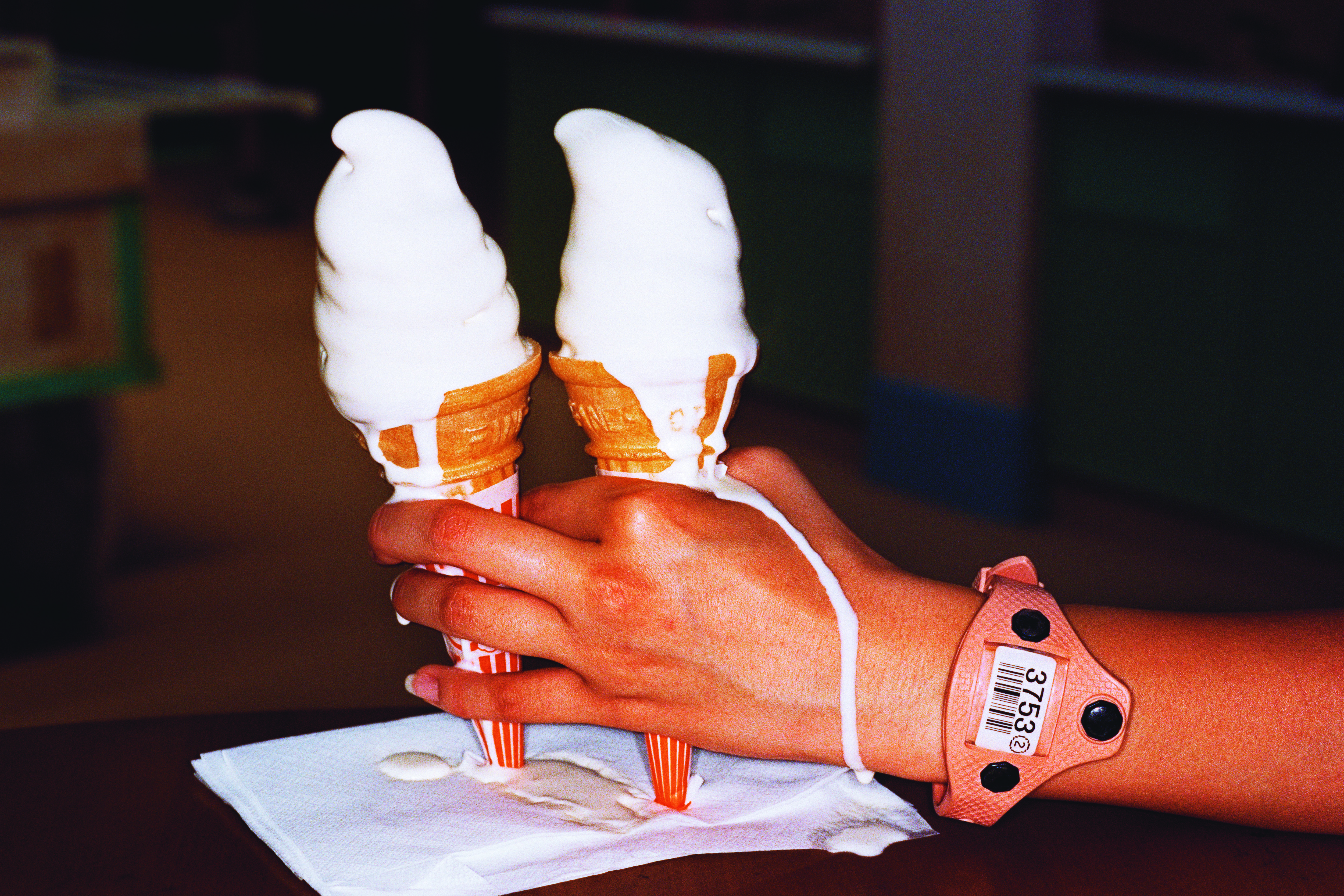 A hand wearing a pink plastic identification wristband holds two soft-serve ice cream cones that are melting and dripping onto a white napkin.