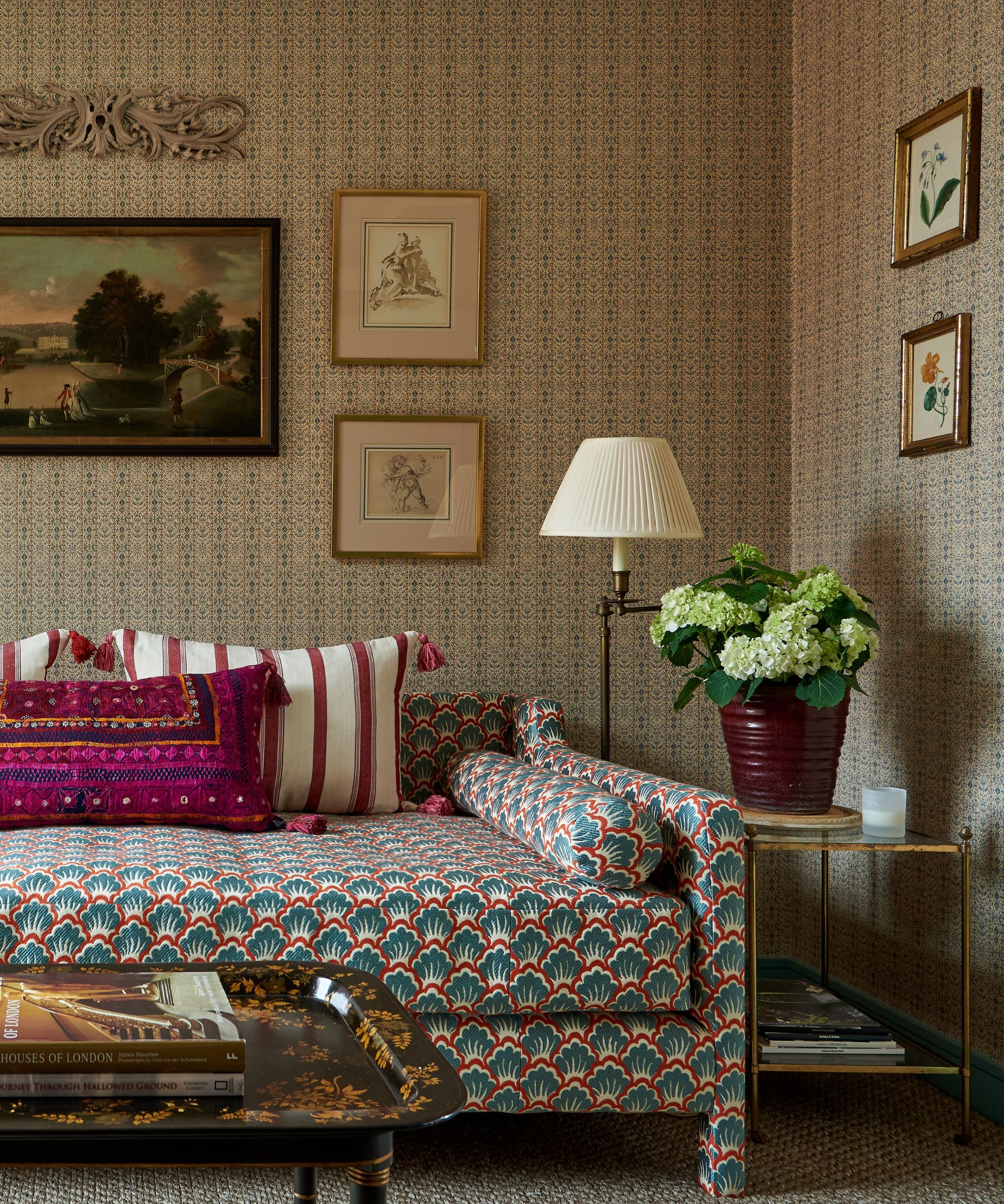 Traditional living room with vintage wallpaper, floral daybed, maroon striped cushion and Indian pink cushion, metal side table with potted hydrangeas, floor lamp and antique coffee table with books
