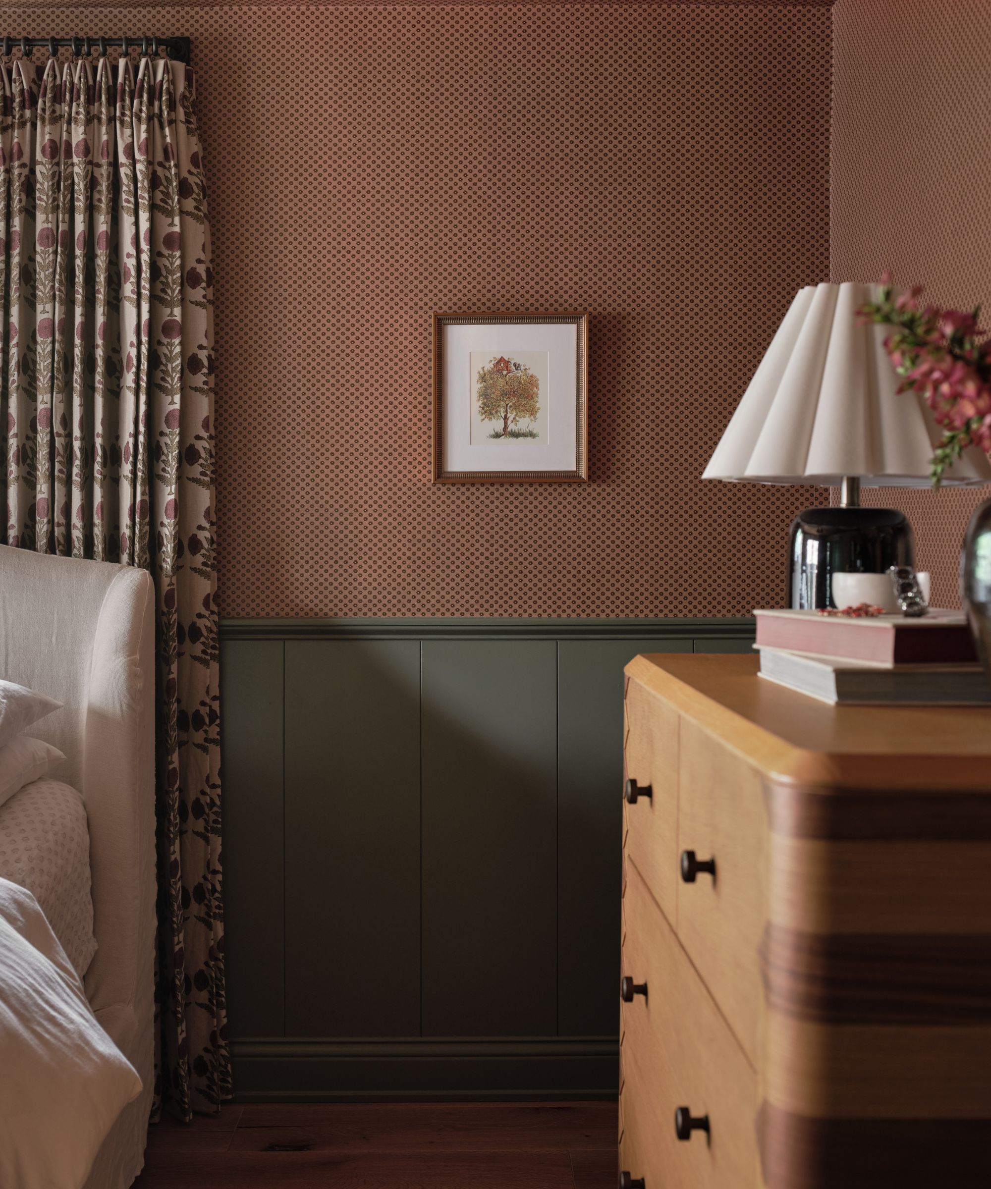 Bedroom with plaster pink wallpaper and a small printed frame, wooden chest of drawers with lamp and books on top and a cream bed with a floral curtain behind it
