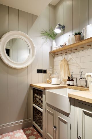 A utility room in neutral shades featuring useful hooks above a large sink, basket storage and a circular mirror